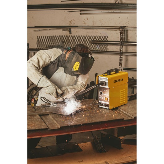 Photo of STANLEY 10880 welding machine in use, operator welding metal in workshop, yellow machine visible on table.