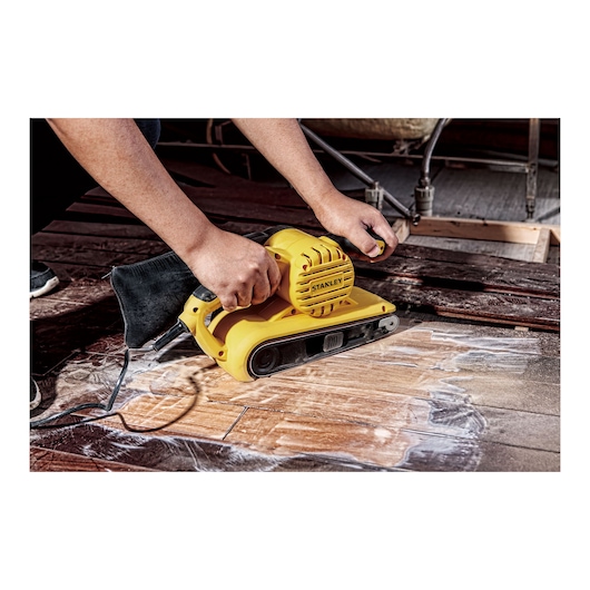STANLEY SB90-B2C belt sander in use on wooden flooring, close-up showing dust bag and ergonomic handle.