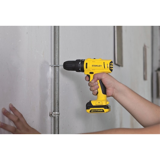 Stanley SCH121S2K-B2 cordless drill, yellow and black, in use drilling metal pipe against wall.