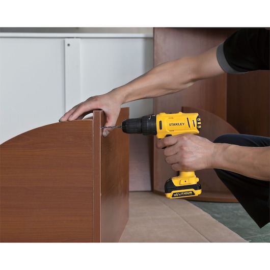 STANLEY SCH121S2K-B2 cordless drill in use drilling into brown wooden furniture, side angle.