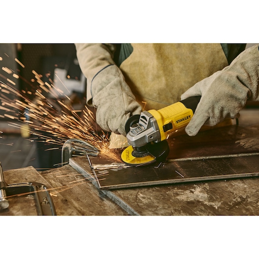 STANLEY stgs9115 angle grinder with yellow handle, sparks flying, used on metal sheet in workshop.