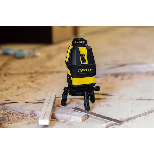 STANLEY STHT77521 laser level on tripod, black and yellow, shown on a workshop floor with wood pieces.