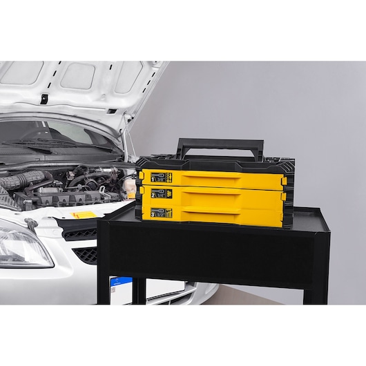 STANLEY STMT17993-LA yellow and black tool organizer box with handle, shown on a workshop cart near a car engine, side angle.
