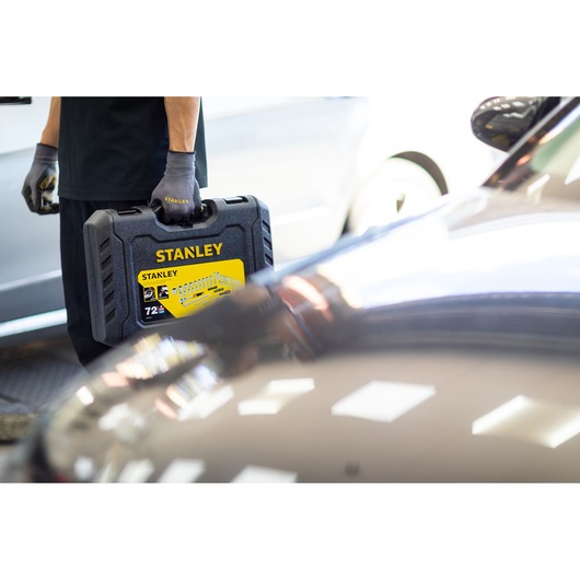 Photo of STANLEY STMT82832-1 tool set case with yellow label, carried by person with gloves near vehicles.