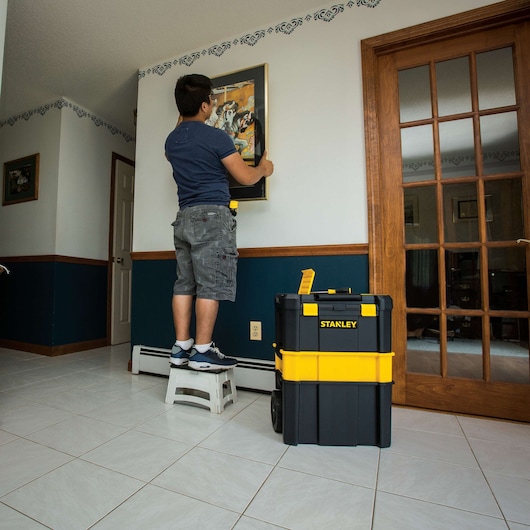 Photo of STANLEY STST18631 black and yellow rolling tool box in a home interior near a person hanging a picture.