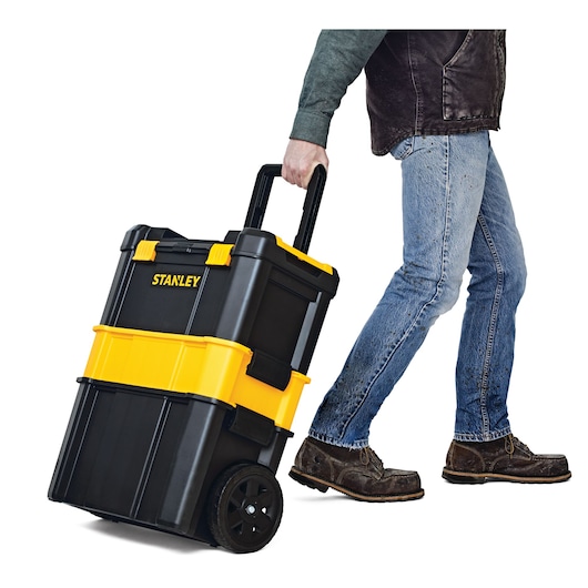 STANLEY STST18631 rolling tool box in black and yellow, shown being pulled by a person, side view.