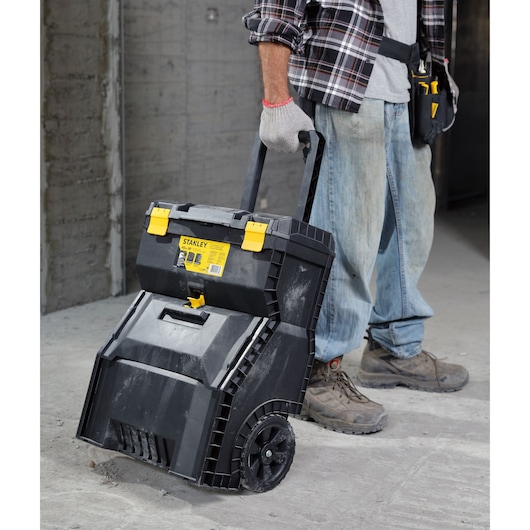 STANLEY STST18800 rolling tool box with black body, yellow latches, and wheels, shown on a construction site floor.