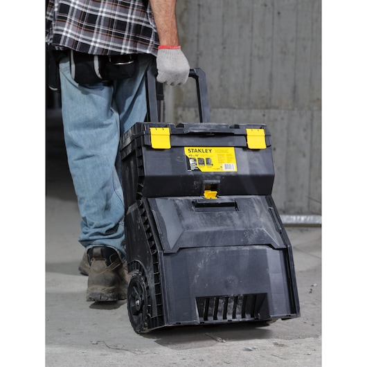 STANLEY STST18800 rolling tool box with yellow latches and handle, being pulled by person in work clothes.