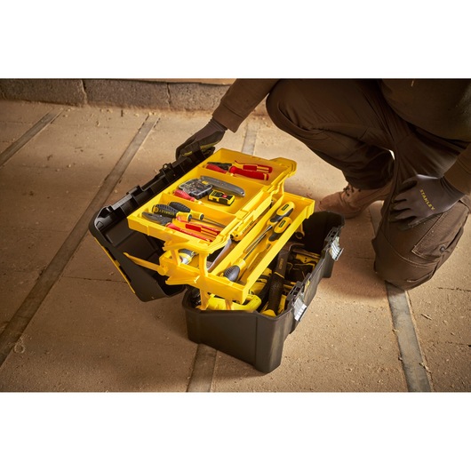 STANLEY STST83397-1 black and yellow tool box with open trays showing assorted hand tools on workshop floor.