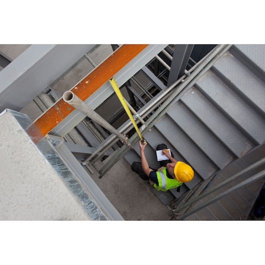 STANLEY XTHT0-33504 tape measure used by worker on stairs at construction site, viewed from above.
